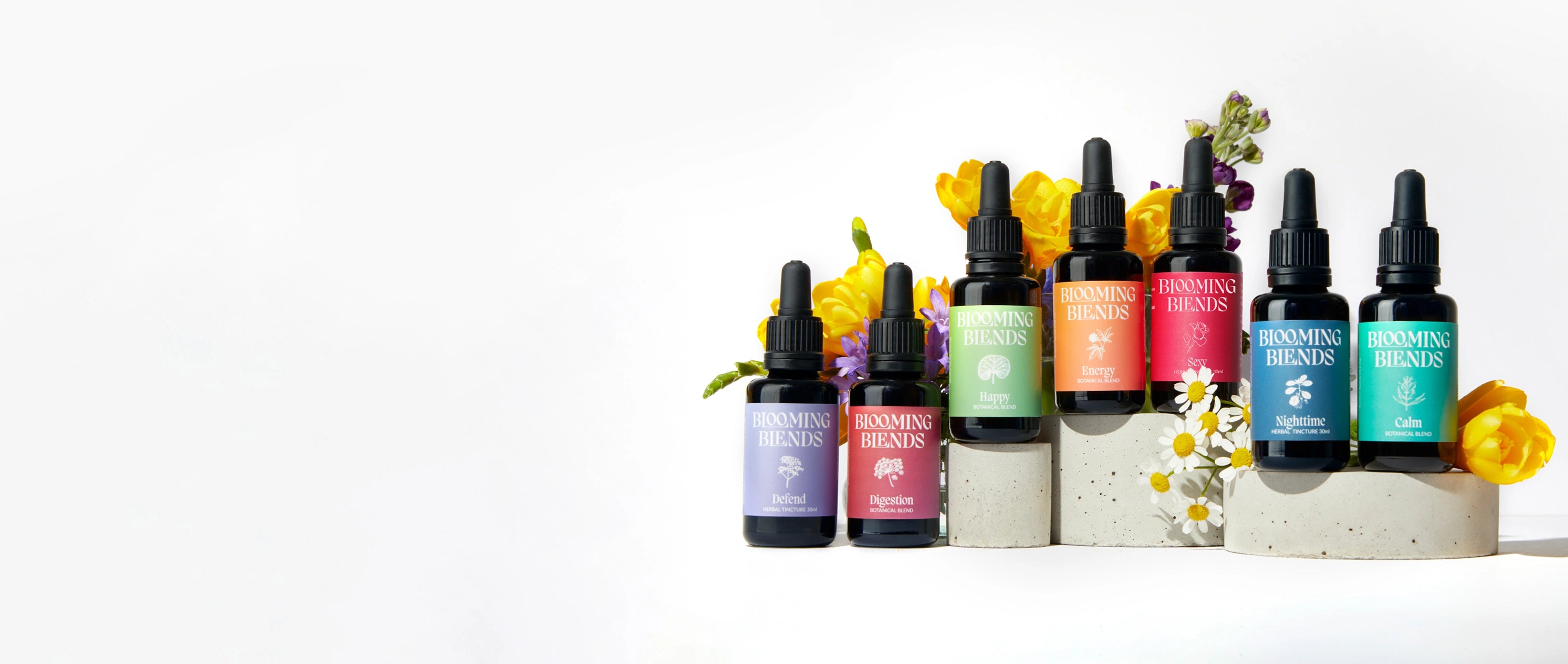 Blooming Blends herbal tincture bottles arranged with fresh flowers on a white background.