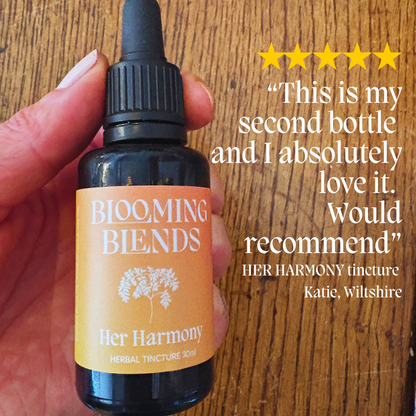 Bottle of Blooming Blends Her Harmony tincture held by a hand on a wooden surface with a five-star rating and review text. Google review - support female hormone balance with traditional herbs linked to supporting the menopause
